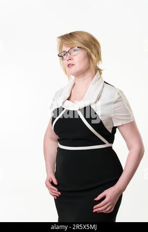 Eine seriöse, selbstbewusste Geschäftsfrau in Brille, gut gekleidet in schwarz-weißem Kleid, die wegschaut. Reife blonde Frauen, die ruhig posieren und die Arme unten halten Stockfoto