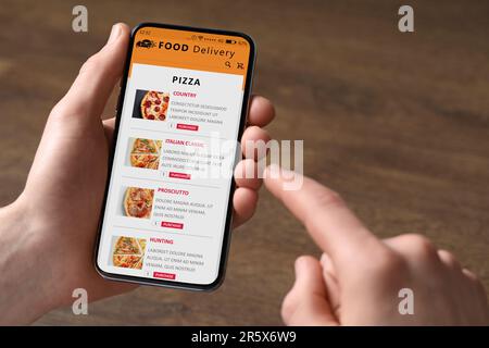 Lebensmittellieferservice. Mann, der ein Gericht aus der Speisekarte vor Ort auswählt, mit Smartphone am Holztisch, Nahaufnahme Stockfoto