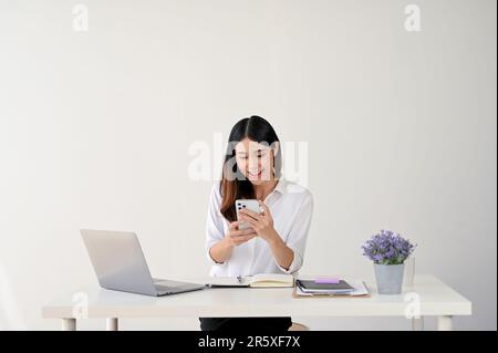 Aufgeregte und fröhliche junge asiatische Büroangestellte, die auf ihren Telefonbildschirm blickt, während sie an ihrem Schreibtisch sitzt. Feiern, jubeln, gute Nachrichten, Gewinn Stockfoto