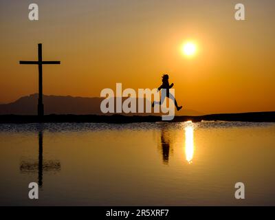 Silhouette einer Frau, die vor Freude neben einem Gipfelkreuz bei Sonnenuntergang springt, Reflexion im Wasser, Trattberg, Bad Vigaun, Land Salzburg, Österreich Stockfoto