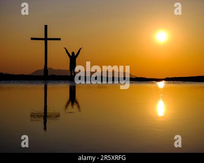 Silhouette einer Frau mit ausgestreckten Armen neben einem Kreuz auf einem Gipfel bei Sonnenuntergang, Reflexion im Wasser, Trattberg, Bad Vigaun, Land Salzburg Stockfoto