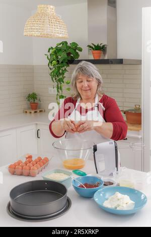 Ältere weißhaarige Frau in Schürze, die Eier knackt, um in ihrer Küche einen Kuchen zu backen Stockfoto
