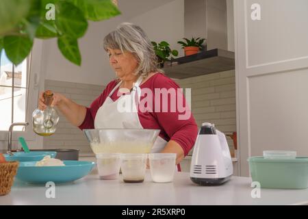 Eine ältere weißhaarige Frau kocht in einer weißen Schürze und mischt Zutaten, um einen Erdbeerkuchen zu machen Stockfoto