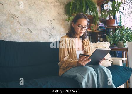 Ich Kriege meinen Schuss, ohne vom Sofa aufzustehen. Eine junge Frau, die zu Hause ein digitales Tablet und eine Kreditkarte auf dem Sofa benutzt. Stockfoto