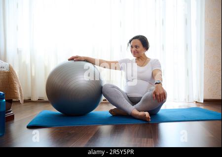 Schöne schwangere Frau, die im Lotus sitzt, posiert auf Yoga-Matte und übt pränatale Dehnung mit Fitnessball zu Hause Stockfoto