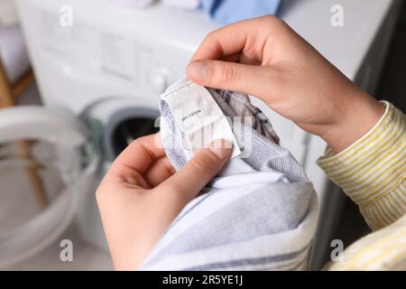 Frau liest Kleidungsetikett mit Pflegesymbolen und Materialinhalt auf Hemd in der Nähe der Waschmaschine, Nahaufnahme Stockfoto