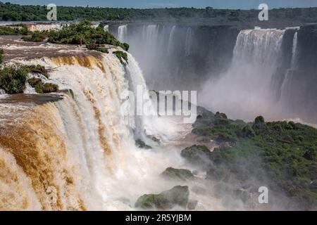 Blick auf die mächtigen Iguazu-Fälle in der Nähe von Devil's Throat mit braunem und weißem Wasser in üppiger grüner Vegetation Stockfoto