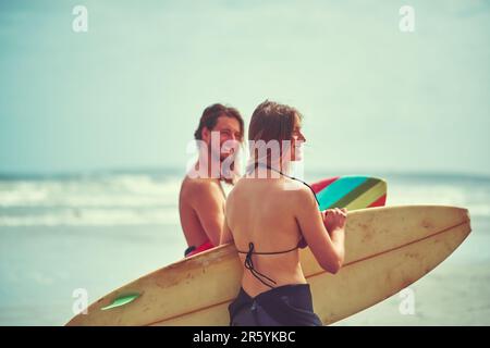 Lasst uns in die Wellen gehen. Ein junges Paar, das mit seinen Surfbrettern am Strand spaziert. Stockfoto