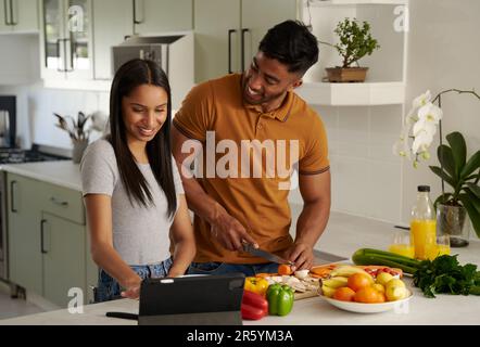 Glückliches junges birassisches Paar in legerer Kleidung, das in der Küche neben einem digitalen Tablet Essen zubereitet Stockfoto