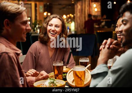 Fröhliche junge, multirassische Gruppe von Freunden in legerer Kleidung, die lachen und ein Abendessen an der Bar genießen Stockfoto