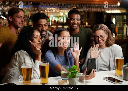 Fröhliche, junge, multiethnische Gruppe von Freunden in legerer Kleidung, die Videoanrufe mit dem Mobiltelefon an der Bar führen Stockfoto