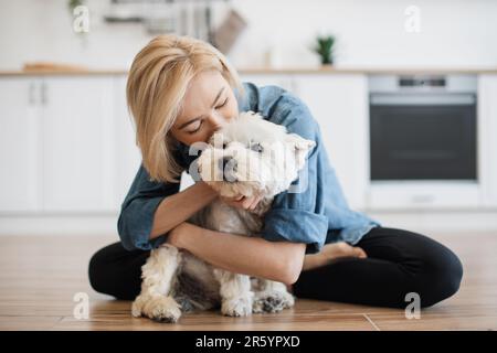 Loyaler West Highland White Terrier in der Umarmung erwachsener Frauen in einem modernen Apartment. Liebende Tierbesitzerin küsst ihre pelzige beste Freundin und genießt dabei die tägliche Interaktion mit dem Hund. Stockfoto