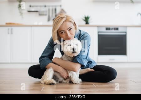 Loyaler West Highland White Terrier in der Umarmung erwachsener Frauen in einem modernen Apartment. Liebende Tierbesitzerin küsst ihre pelzige beste Freundin und genießt dabei die tägliche Interaktion mit dem Hund. Stockfoto