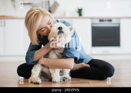 Loyaler West Highland White Terrier in der Umarmung erwachsener Frauen in einem modernen Apartment. Liebende Tierbesitzerin küsst ihre pelzige beste Freundin und genießt dabei die tägliche Interaktion mit dem Hund. Stockfoto