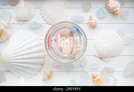 Von oben sehen Sie die hausgemachte Unterwasserkerze aus klarem Gel mit Muschel in einem durchsichtigen Becher. Wunderschöne Kerze auf weißem Holztablett mit Meeresglas und Muscheln. Stockfoto