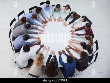 Eine Gruppe junger Menschen, die mit ausgestreckten Armen an einem runden Tisch sitzen Stockfoto