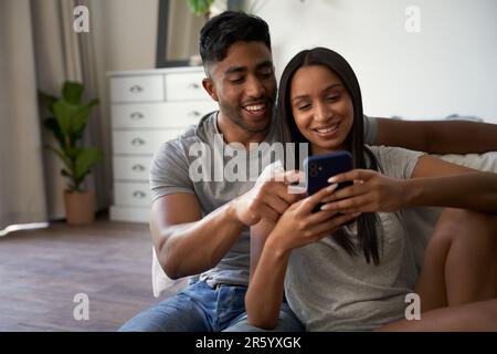 Glückliches junges birassisches Paar in lässiger Kleidung, das zu Hause ein Handy im Schlafzimmer benutzt Stockfoto