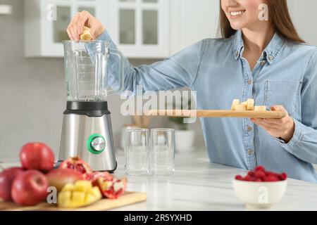 Wunderschöne junge Frau, die Banane in den Mixer gibt, um leckeren Smoothie in der Küche zu genießen Stockfoto