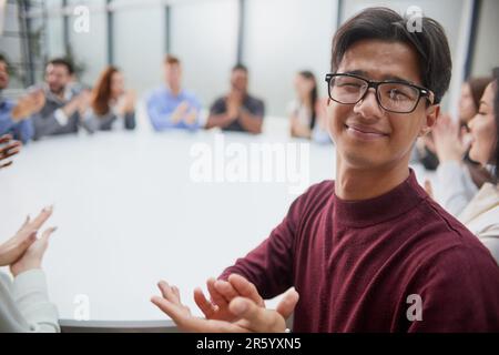 Junger, gutaussehender Geschäftsmann mit Brille, der vor dem Hintergrund seiner Kollegen für die Kamera posiert Stockfoto