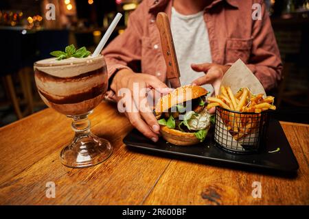 Nahaufnahme eines jungen Weißen in legerer Kleidung mit Burger und Chips auf dem Tisch im Restaurant Stockfoto