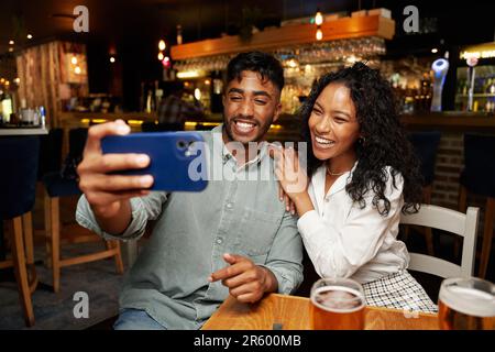Ein junges, multiethnisches Paar in legerer Kleidung, das lächelt und Selfie mit dem Handy an der Bar macht Stockfoto