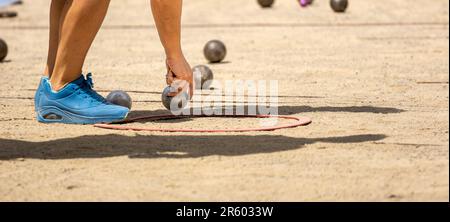 Die Hand einer jungen Sportlerin in Sportschuhen, die sich einen Petanque-Ball aus Metall fängt, um sich beim Qualifikationsspiel zu messen und an einem sonnigen Tag auf einem Sandy Petanque-Ball zu spielen Stockfoto