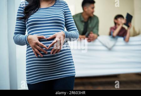 Sie hat nur bedingungslose Liebe für ihre Familie. Eine schwangere Frau, die zu Hause ein Herz mit den Händen auf dem Bauch formt. Stockfoto