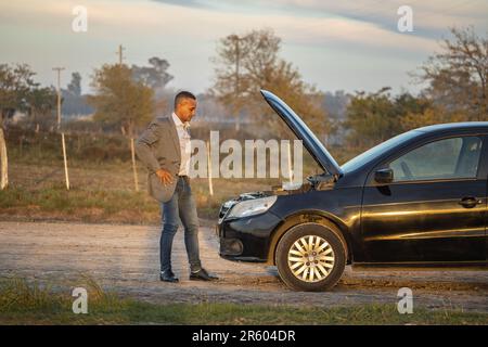 Ein junger Latino in einem Anzug, der den Motor seines kaputten Autos am Straßenrand ansieht. Stockfoto