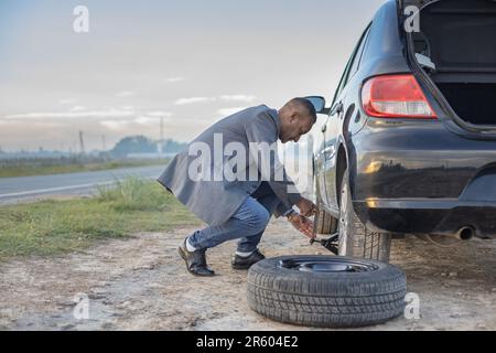 Ein Latino-Mann im Anzug wechselt einen Platten Reifen an seinem Auto am Straßenrand mit Kopierraum. Stockfoto