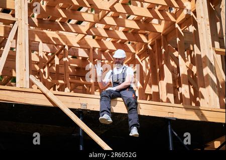Der männliche Architekt auf der Baustelle des holzgerahmten Hauses gezeichnet das OK-Zeichen. Das Konzept bezieht sich auf den modernen ökologischen Bau. Stockfoto