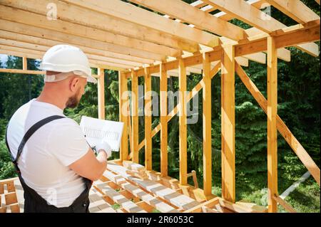 Baumeister, der ein zweigeschossiges Holzgebäude in der Nähe des Waldes errichtet. Rückansicht eines Mannes in Arbeitsausrüstung und Schutzhelm untersucht den Bauplan. Konzept moderner und nachhaltiger Baumethoden. Stockfoto