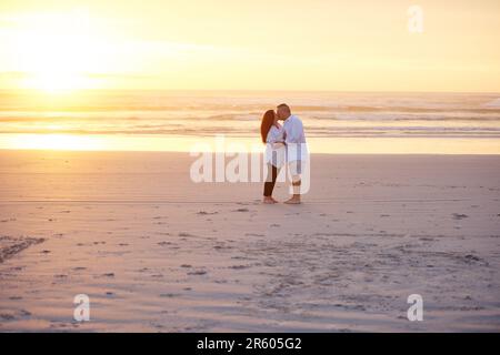 Nichts sagt Romantik besser als ein Kuss bei Sonnenuntergang. Ein reifes Paar, das sich am Strand küsst. Stockfoto