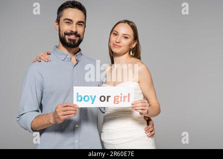 Eine positive und trendige schwangere Frau, die eine Karte mit Buchstaben von Jungen oder Mädchen hält und den stilvollen Ehemann während des Geschlechts umarmt, zeigt eine Überraschungsparty isoliert auf Stockfoto