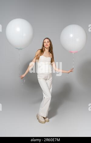 Eine trendige und schwangere Frau blickt während der Feier weg, während sie weiße festliche Ballons hält, und das Geschlecht zeigt eine Überraschungsparty auf Grau Stockfoto