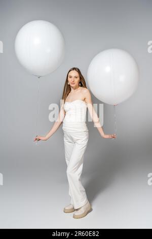 Die ganze Länge einer trendigen und blonden schwangeren Frau, die weiße Ballons hält und während des Geschlechts in die Kamera schaut, enthüllt eine Überraschungsparty auf grauem Hintergrund Stockfoto