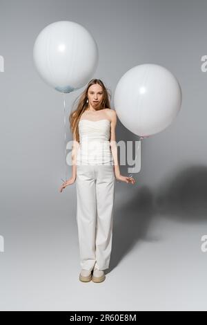 Die ganze Länge voller stilvoller und schockierter Überraschungsparty auf Grau zeigt die Mutter, die in die Kamera schaut, während sie während des Geschlechts weiße festliche Ballons in der Hand hält Stockfoto