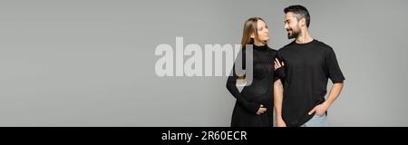 Stilvolle und schwangere Frau in schwarzem Kleid, die den Arm eines lächelnden bärtigen Ehemannes berührt und isoliert auf grauen, neuen Anfängen und Elternschaft zusammensteht Stockfoto