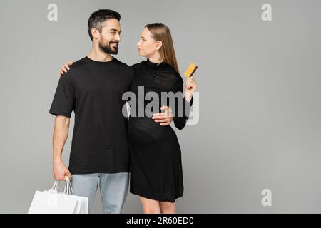 Lächelnder bärtiger Mann mit Einkaufstaschen und stilvolle schwangere Frau in schwarzem Kleid mit Kreditkarte isoliert auf Grau, Neuanfang und p Stockfoto