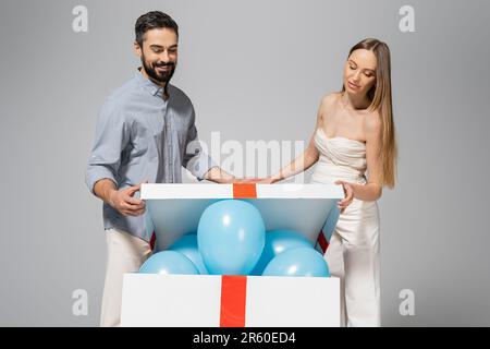 Fröhliche und trendige Eltern, die eine große Geschenkbox mit festlichen blauen Luftballons öffnen, während das Geschlecht die Überraschungsparty und die Feier isoliert auf der enthüllt Stockfoto