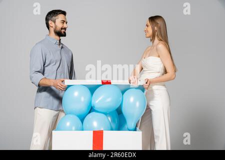 Fröhliche und stilvolle schwangere Frau, die ihren Ehemann ansieht, während sie eine große Geschenkbox mit festlichen blauen Ballons öffnet, während das Geschlecht die Überraschungsparty isola enthüllt Stockfoto