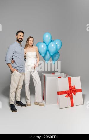 Die ganze Länge voller trendiger und fröhlicher erwartender Eltern umarmt sich, während sie neben blauen Ballons stehen und eine große Geschenkbox, während Gender Reveal Surpri Stockfoto