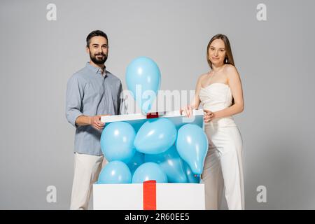 Positiv und stilvoll erwartende Eltern schauen in die Kamera, während sie eine große Geschenkbox mit blauen Ballons öffnen, während sie eine Überraschungsparty und Feier aufdecken Stockfoto