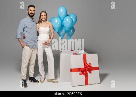 Die ganze Länge voller fröhlicher und trendiger Eltern, die sich umarmen und in die Kamera schauen, während sie in der Nähe einer großen Geschenkbox und festlicher blauer Ballons während g stehen Stockfoto