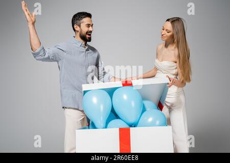 Ein bärtiger Mann, der bei einer stilvollen schwangeren Frau verliert, öffnet eine große Geschenkbox mit blauen Ballons während der Gender-Show Überraschungsparty und Feier-Isolat Stockfoto