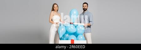 Lächelnde und trendige Eltern erwarten, dass sie auf die Kamera schauen, während sie die Moorschachtel mit festlichen blauen Ballons öffnen, während das Geschlecht die Überraschungsparty isola enthüllt Stockfoto
