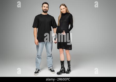 Eine stilvolle, blonde, schwangere Frau in schwarzem Kleid, die Hand eines bärtigen Ehemannes hält und auf grauen Hintergrund in die Kamera schaut, Concept Stockfoto