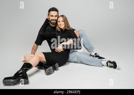 Eine fröhliche, modische und schwangere Frau in schwarzem Kleid und Stiefeln, die in die Kamera schaut und ihren Ehemann umarmt, während sie auf grauem Hintergrund sitzt, ein Neuanfang Stockfoto
