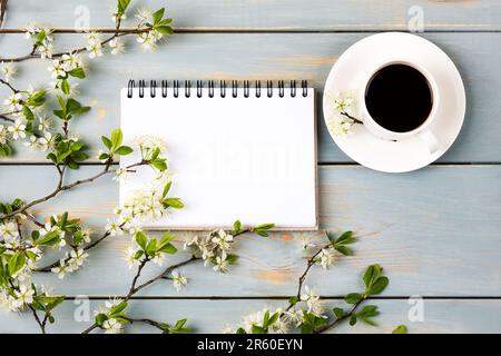 Künstlerische Komposition mit offenem leeren Notizblock, einer Tasse schwarzen Kaffees, einem Zweig weißer Kirschblüten auf einem blauen Holztisch. Festliches Büro Desktop Conc Stockfoto