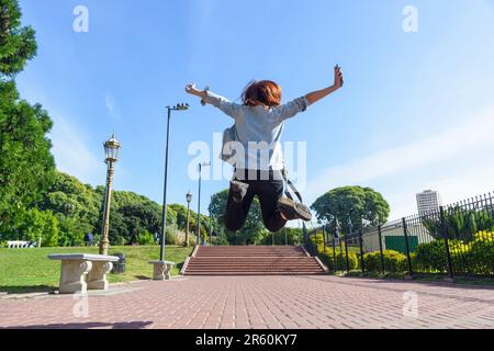 Rückansicht einer jungen Frau, die mit offenen Beinen und Armen in den Park springt, sie trägt eine jeansjacke und -Hose, rote Haare und den Himmel hinten Stockfoto