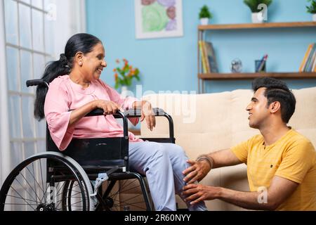 Indischer erwachsener Sohn, der auf dem Schoß der Seniormutter schläft, indem er die Beine drückt, während die Mutter auf dem Rollstuhl sitzt - Konzept von Fürsorge, Familienunterstützung und Stockfoto